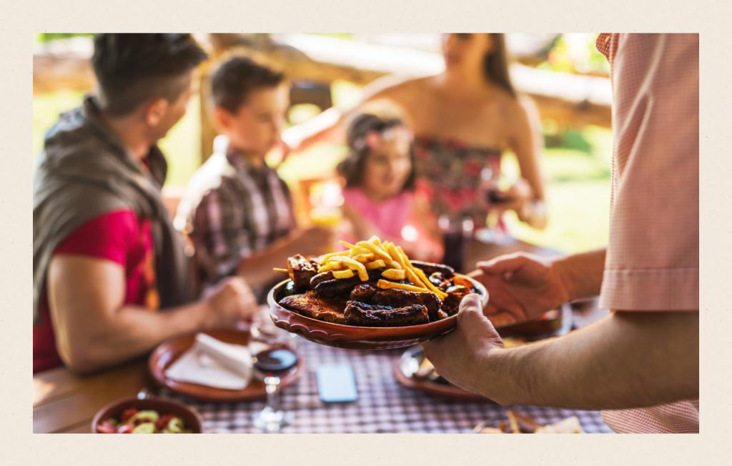 A man holding BBQ food on a plate with a happy family in the background.