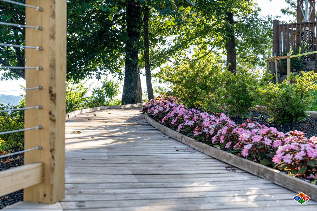 Wooden boardwalk at Cielito Lindo edged with pink impatiens, leading toward the deck under the trees
